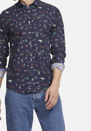 Námořnická košile s barevnými vzory drobných postav, stromů a lodí nošená s modrými džínami, rukávy vyhrnuté.