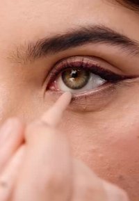 Close-up van een oog met een warme bruine iris met groene vlekken, omrand door donkere eyeliner, terwijl een vinger make-up aanbrengt nabij de waterlijn.