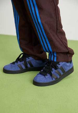 Zapatillas de gamuza azul con rayas y cordones negros. La persona lleva pantalones marrones con estampado y rayas azules laterales. Superficie verde debajo.