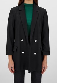 Blazer nero doppiopetto con grandi bottoni bianchi, rever sagomati e maniche arrotolate, indossato sopra un dolcevita verde a coste.