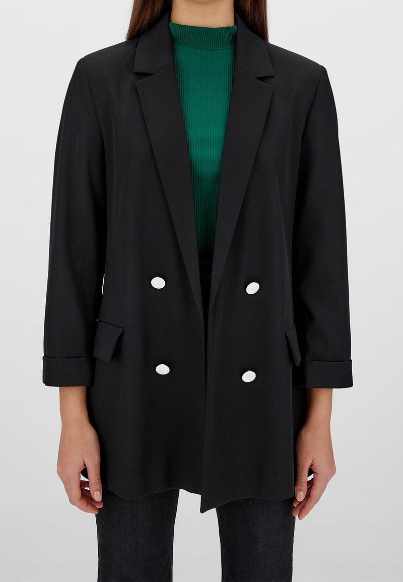 Blazer nero doppiopetto con grandi bottoni bianchi, rever sagomati e maniche arrotolate, indossato sopra un dolcevita verde a coste.