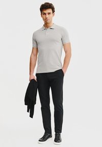 Jasnoszara koszulka polo z krótkim rękawem, w zestawie z czarnymi spodniami slim fit i czarnymi trampkami, trzymana czarna kurtka pod pachą.