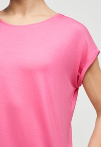 Κοντινό πλάνο ενός ατόμου που φοράει φωτεινό ροζ κοντομάνικο πουκάμισο με στρογγυλή λαιμόκοψη, μπροστά σε απλό ανοιχτό φόντο.