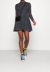 Frau in einem schwarzen, blumigen Langarmkleid, mit einer schwarzen Umhängetasche, neongelben Socken und schwarzen High-Top-Sneakern, die nach vorne läuft.