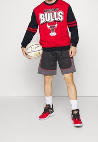 Sweatshirt rouge et noir des Chicago Bulls avec logo, short de basket en mesh gris avec accents rouges, et baskets rouges et noires, tenant un ballon de basket.