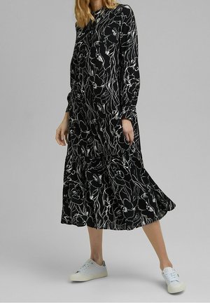 Femme portant une robe midi noire avec de grands motifs floraux blancs et des baskets blanches, debout devant un fond gris uni.