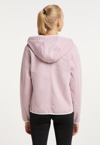 Világos rózsaszín kapucnis pulóver, zsinóros kapucnival, látható varrási részletekkel és fehér díszítéssel az alján. Fekete leggings fölött viselve.