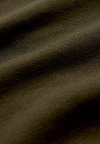 Tmavozelená látka s jemnou textúrou, s jemným tkaním. Materiál pôsobí hladko s miernymi vlnami a záhybmi.