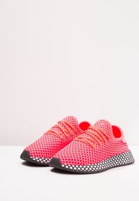 Klarröda träningsskor med ett meshinstrument, med ett triangulärt mönster, texturerade snören och en sål med svart-vitt mönster.