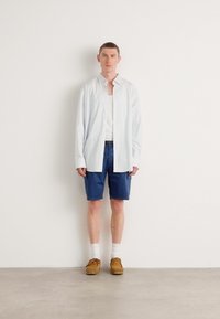 Chemise bleu clair à boutons, t-shirt blanc en dessous, shorts navy, chaussures marron, chaussettes blanches. Le mannequin se tient devant un mur blanc uni.