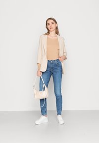 Blazer xadrez bege, top canelado bege, jeans skinny azuis, ténis brancos e uma pequena mala creme com uma alça trançada. Outfit minimalista e moderno.