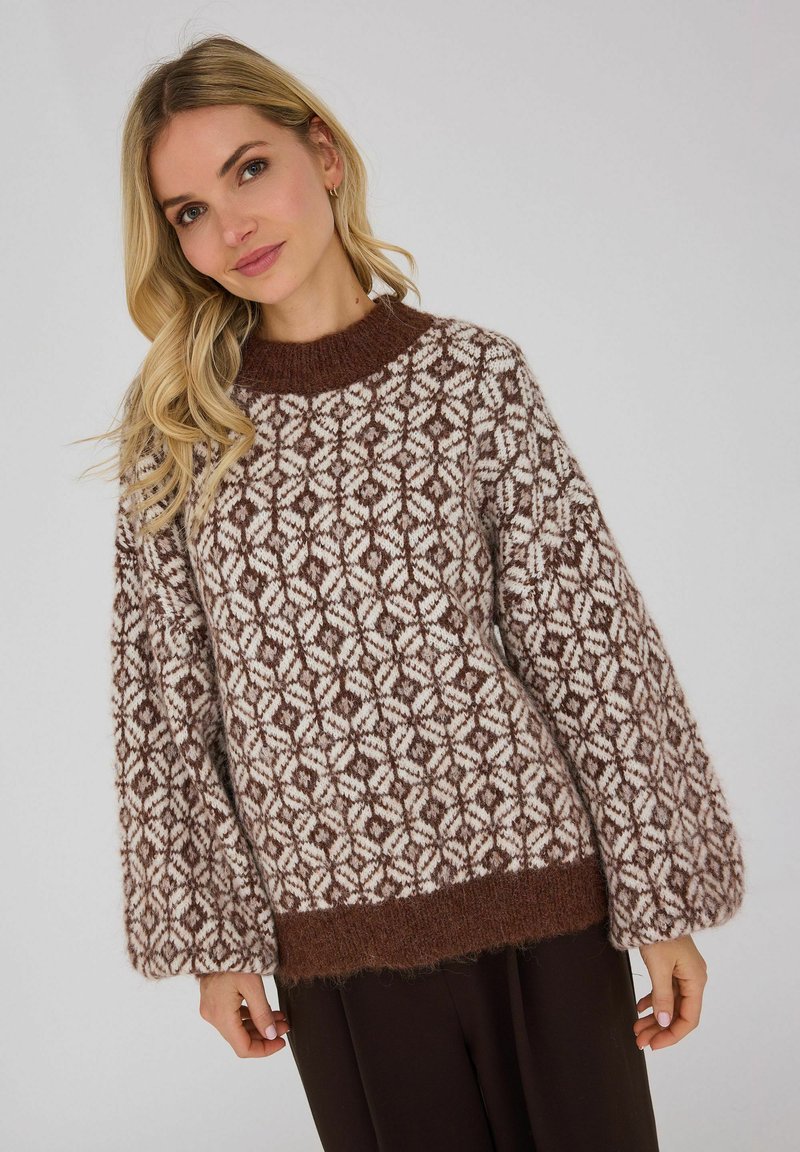 Femme blonde portant un pull en maille marron et blanc à motifs avec manches ballon et un pantalon marron foncé sur un fond uni.