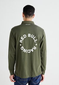 Olivno zelena polo majica z dolgimi rokavi in zaobljenim robom, na hrbtni strani pa se nahaja velika bela natisnjena značka, ki pravi "RED BULL RACING."