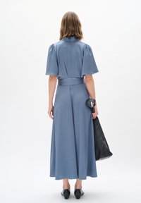 Langes, hellblaues Kleid mit kurzen, puffärmeligen Ärmeln, einem taillierten Gürtel und einem fließenden Saum. Getragen mit einer schwarzen Ledertasche und Schuhen.