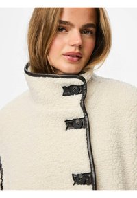 Kremfarget, myk fleecejakke med høy krage, pyntet med mørkebrune skinnkanter og tre mørke knapper. Myk tekstur, minimalistisk design.