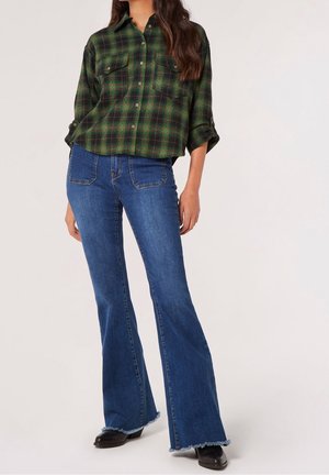 Woman wearing green and black plaid button-up shirt with rolled sleeves, blue flared jeans with front pockets, and black shoes.