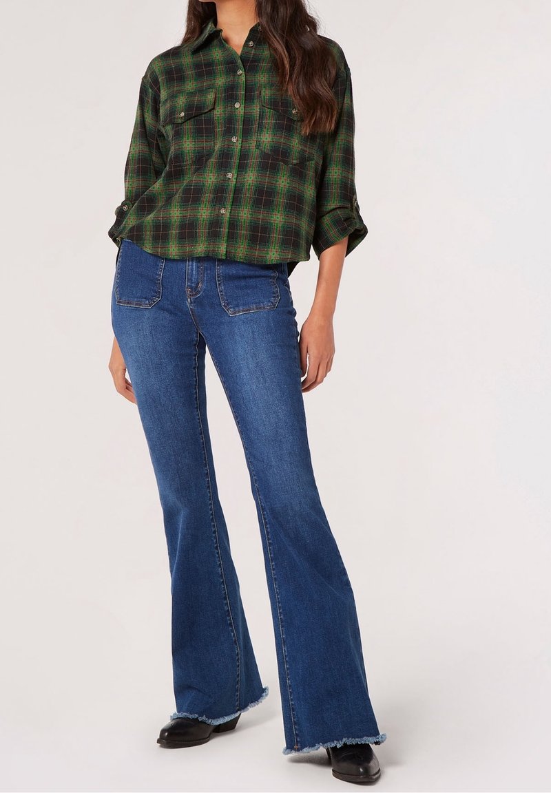 Femme portant une chemise à carreaux verte et noire à boutons avec manches retroussées, un jean bleu évasé avec poches avant, et des chaussures noires.
