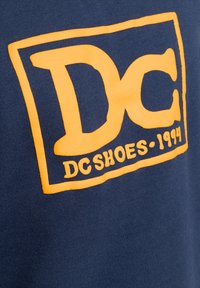 Navy blue sweatshirt featuring a large, orange DC Shoes logo from 1994, with a soft texture and bold, graphic design against the fabric.