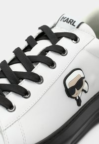 Białe skórzane sneakersy z czarnymi sznurowadłami i oczkami. Posiadają naszywkę z kreskówkową postacią w okularach przeciwsłonecznych po boku oraz logo na języku.