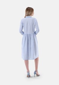 Himmelblaues Hemdkleid mit gerafftem Taillenzug und langen Ärmeln. Glatte Textur, knielang, kombiniert mit hellblauen hohen Schuhen.