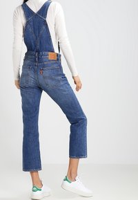 Blue denim overalls featuring a bib front, adjustable straps, and a relaxed fit. Worn with white sneakers accented in green.