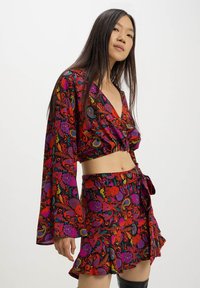 Completo due pezzi in stile uncinetto caratterizzato da un crop top a maniche lunghe con scollatura a V profonda e una mini gonna avvolgente. Vivace motivo floreale in rosso, viola e verde.