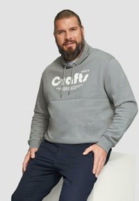 Szary sweter z wysokim kołnierzem, z białym nadrukiem. Gładka tekstura, z regulowanymi sznurkami i ściągaczami na rękawach.