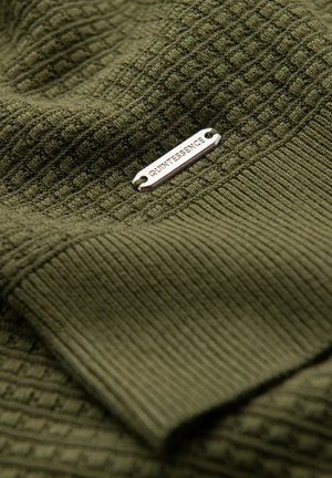 Olijfgroen stof met een gestructureerde brei- en ribpatronen. Bevat een zilveren metalen label met de tekst "QUINTESSENCE." Zacht, gelaagd uiterlijk.