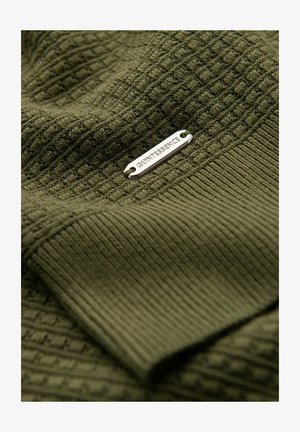 Olivově zelená tkanina s texturovaným pletením a žebrovanými vzory. Obsahuje stříbrnou kovovou nálepku s nápisem "QUINTESSENCE." Měkký, vrstvený vzhled.