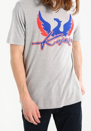 Personne portant un t-shirt gris avec un grand graphisme de phénix bleu et rouge et le mot « Kosmic » sur la poitrine, associé à un pantalon foncé.