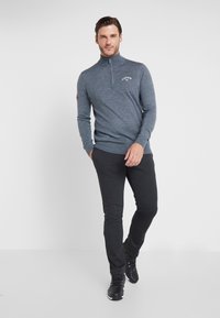Pullover cinza em malha com fecho de meio zip e um pequeno logo no peito, combinado com calças pretas de corte slim e botas pretas de atacador.