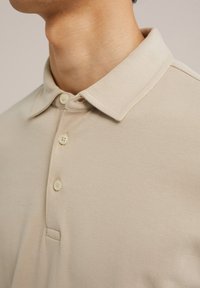 Bež polo majica iz gladkega materiala, z nazobčanim ovratnikom, tri gumbnimi zatiči in subtilnimi detajli šivanja.