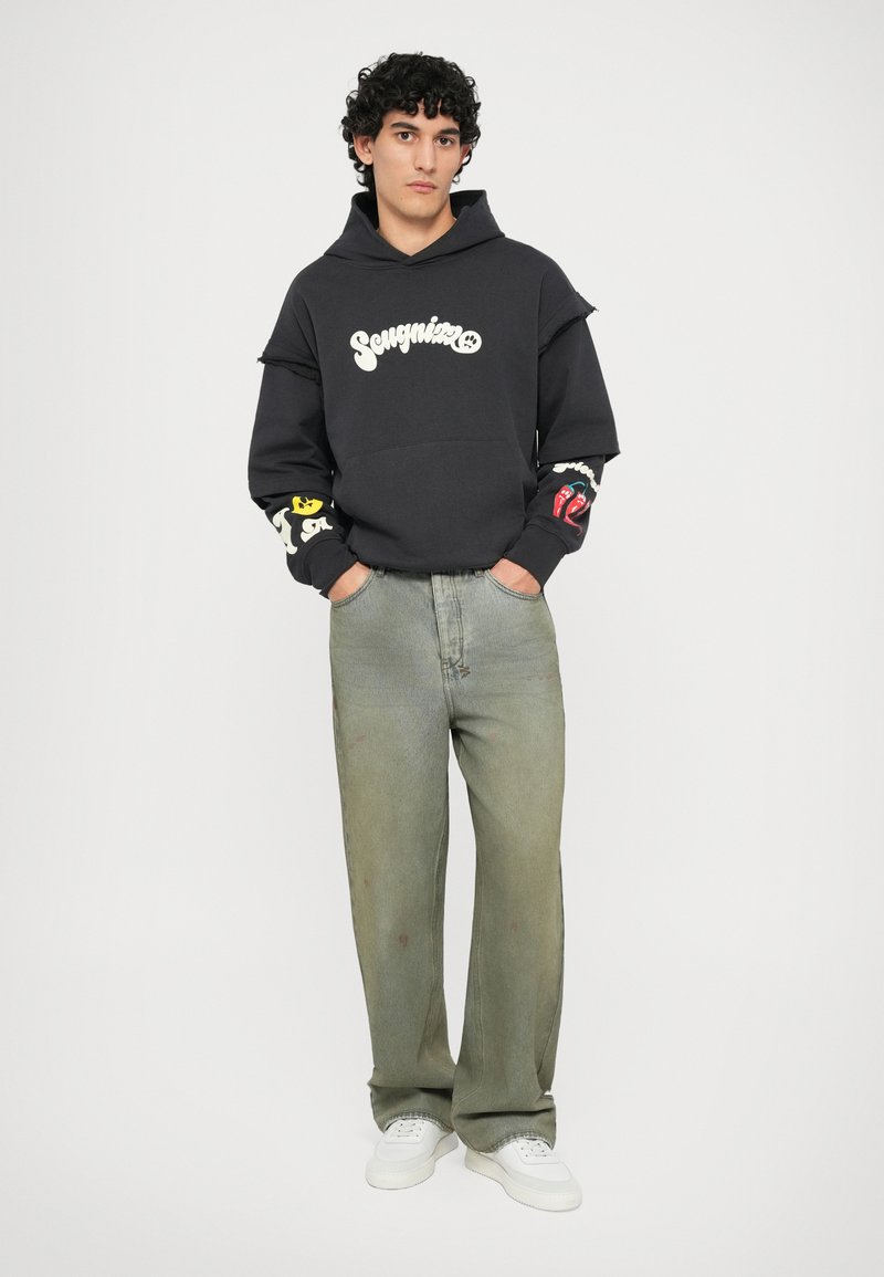 Felpa nera con logo "Songni", grafiche colorate sulle maniche; abbinata a jeans verde chiaro, a vestibilità larga e scarpe da ginnastica bianche.
