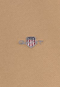 Un tejido de punto beige presenta un pequeño logo de escudo bordado con acentos en azul, rojo y blanco, y la palabra "GANT" en plata.