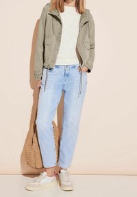 Veste légère olive avec capuche, pull crème, jean bleu clair et sneakers blanches avec accent rose, tenant un sac en osier tan.
