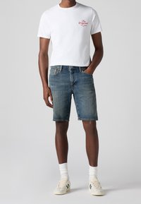 T-shirt blanc à manches courtes avec un motif rouge, short en denim bleu et baskets crème avec des rayures vertes, mettant en avant une tenue décontractée.