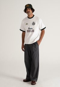 Bărbat îmbrăcat cu o șapcă neagră, un tricou alb cu grafică, pantaloni largi cu dungi subtiri și pantofi maro, stând pe un fundal deschis simplu.