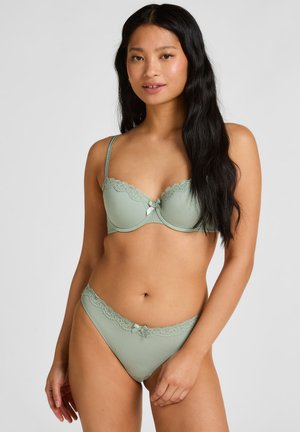 Woman wearing matching light green ribbed lingerie set with lace trim and small bows, standing against a plain light background.