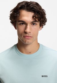 Νέος άνδρας με σγουρά σκούρα μαλλιά, που φοράει ανοιχτό μπλε μπλουζάκι με στρογγυλή λαιμόκοψη και το λογότυπο "BOSS", κοιτάζοντας προς τα εμπρός με φόντο λευκό.