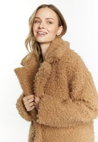 Płaszcz teddy w jasnobrązowym kolorze, z teksturowanej, puszystej tkaniny, z oversize'owym kołnierzem i szerokimi rękawami. Widoczny detal zapięcia z przodu.