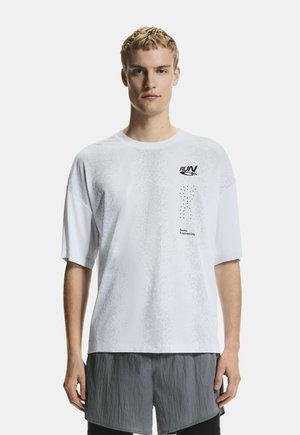 Mladi moški stoji v beli široki majici s kratkimi rokavi z napisom "RUN" in "Swiss Engineering" ter sivih kratkih hlačah.