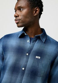Camisa de cuadros azules hecha de una tela de textura suave, con un diseño de botones, un bolsillo y una etiqueta de marca en el pecho.
