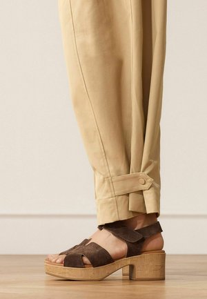Beige taps toelopende broek met enkelbanddetail, gedragen met bruine suède platformsandalen met dikke houten hakken.