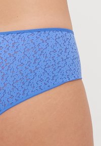 Bas de bikini en dentelle bleu clair avec un motif floral subtil, texture lisse et taille élastique plate. La vue de côté montre une couverture modérée.