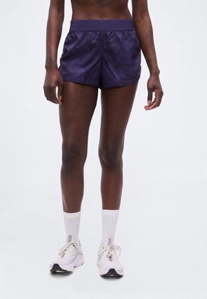 Άτομο που φοράει σκούρα μοβ αθλητικά σορτς, λευκές κάλτσες και λευκά αθλητικά παπούτσια, στέκεται μπροστά σε ένα απλό φόντο.