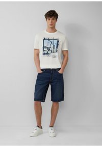Wit T-shirt met korte mouwen en een blauw grafisch ontwerp, gecombineerd met donkere denim shorts en witte sneakers, staand tegen een neutrale achtergrond.