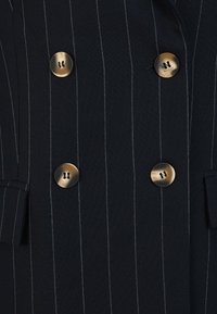 Blazer croisé à rayures marines, avec des boutons dorés et des poches en tissu, présentant une texture lisse et des détails de couture structurée.