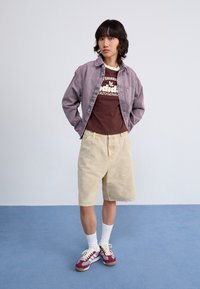 Modell bär en ljuslila denimjacka, brunt grafiskt t-shirt, beige shorts och vinröda sneakers med vita detaljer och strumpor.