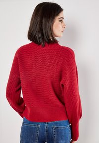 Pull en laine rouge avec un col montant, motif texturé et poignets côtelés. Porté avec un jean bleu, montrant une coupe décontractée et des épaules tombantes.