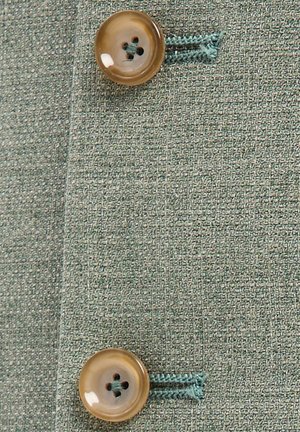 Zwei beige Knöpfe, mit blauem Faden auf strukturiertem grünem Stoff genäht, zeigen Knopflöcher und Stoffdetail.